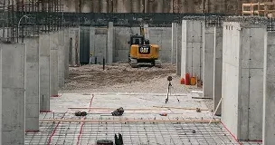 A construction site featuring concrete walls and columns, rebar on the floor, and a yellow professional excavator parked in the background. Various construction materials and orange cones are scattered throughout the area. - Professional Excavators & Construction