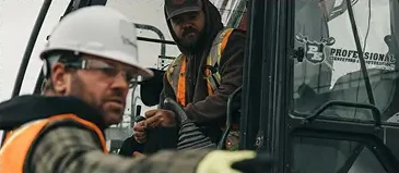 Two professional excavators, dressed in safety vests and hard hats, communicate near heavy construction machinery—one inside the cab and the other pointing outside. - Professional Excavators & Construction
