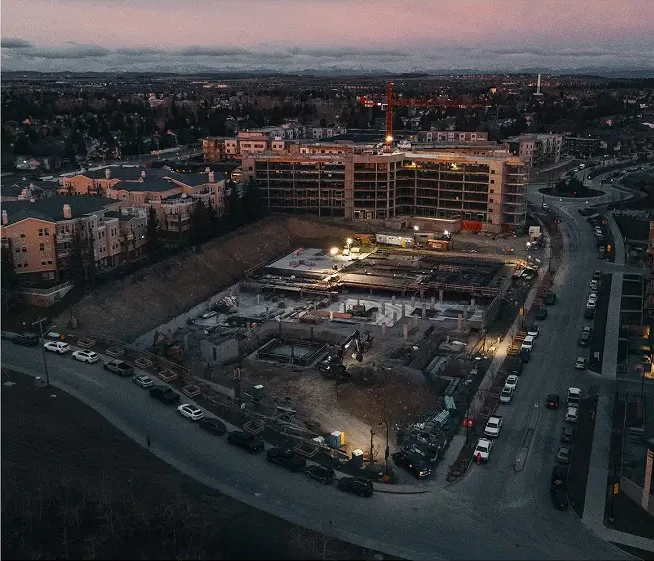 Aerial view of a construction site at dusk, illuminated by work lights and surrounded by parked cars and homes. A large, partially-built structure rises in the background—get in touch with Professional Excavators and Construction for your next project. - Professional Excavators & Construction