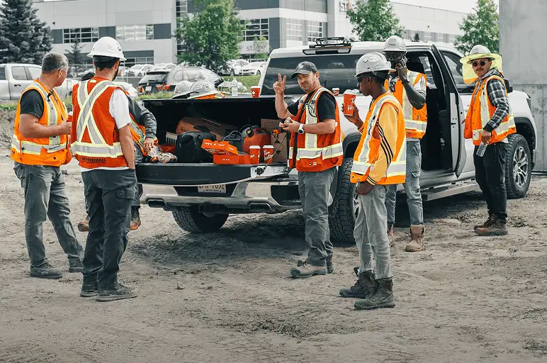 A group of Professional Excavators and Construction workers in safety vests and hard hats gather around the open tailgate of a pickup truck filled with tools at a worksite. Some are talking, and one is waving toward the camera for our About Us page. - Professional Excavators & Construction