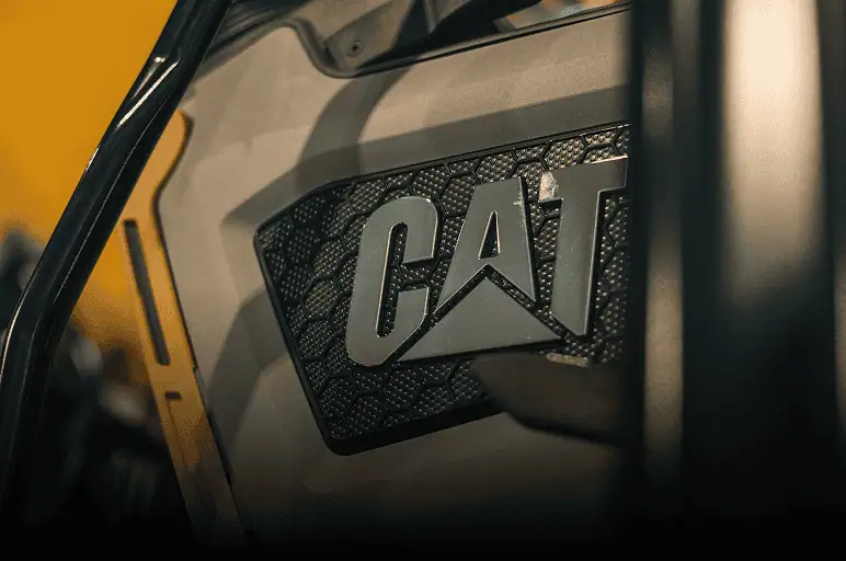 Close-up of a metallic grill with the bold "CAT" logo, part of Caterpillar construction equipment used by Professional Excavators and Construction, set against a warm yellow background and black frame elements. - Professional Excavators & Construction