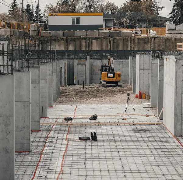 Construction site with concrete columns, rebar, and equipment, featuring a yellow CAT excavator from Professional Excavators and Construction in the center. A temporary building stands in the background, with trees and an overcast sky beyond the area. - Professional Excavators & Construction