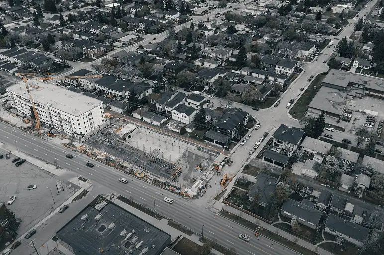 Aerial view of a suburban neighborhood with a Professional Excavators and Construction site near a main road, surrounded by houses, trees, and parked cars on a cloudy day. - Professional Excavators & Construction