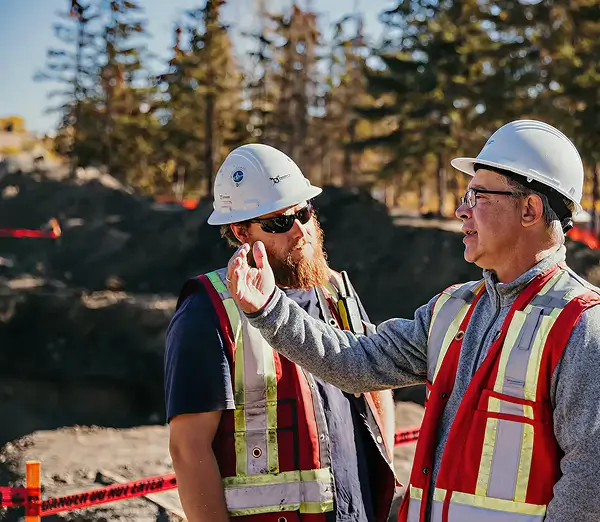 Two construction workers wearing hard hats, safety vests, and sunglasses stand outdoors at a construction site, engaged in conversation. As professional excavators and construction experts, they provide top-quality services with trees and equipment in the background. - Professional Excavators & Construction