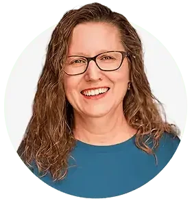 A woman with wavy brown hair and glasses smiles at the camera. She is wearing a blue top and represents the Professional Excavators and Construction team, posed confidently in front of a plain white background. - Professional Excavators & Construction