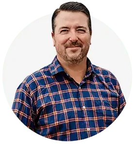 A man with short dark hair and a trimmed beard is smiling, wearing a blue and orange plaid shirt. He’s part of the Professional Excavators and Construction team. The background is plain white. - Professional Excavators & Construction