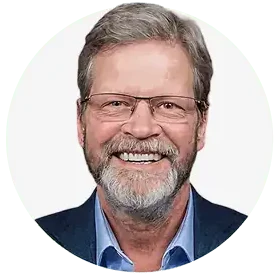 A middle-aged man with gray hair, a beard, and glasses, smiling. He is wearing a blue collared shirt and a dark jacket, posing against a white background—representing the Team at Professional Excavators and Construction. - Professional Excavators & Construction