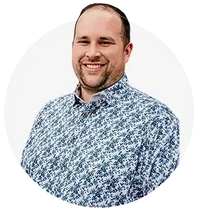 A man with short hair and a trimmed beard smiles at the camera. He is wearing a blue and white floral-patterned button-up shirt, representing the Team at Professional Excavators and Construction, against a plain, light background. - Professional Excavators & Construction