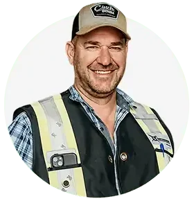 A smiling man wearing a beige cap, plaid shirt, and reflective safety vest stands facing forward, representing the Professional Excavators and Construction team. His vest has a smartphone in the pocket and a visible patch on the chest. Plain white background. - Professional Excavators & Construction