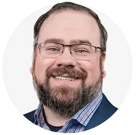 A smiling man with glasses, short dark hair, and a beard, wearing a blue blazer over a plaid shirt, represents the Professional Excavators and Construction team against a white background. - Professional Excavators & Construction