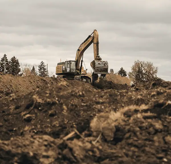 A large excavator from Professional Excavators and Construction is digging and moving dirt on a construction site, with piles of soil in the foreground and trees in the background under a cloudy sky. - Professional Excavators & Construction