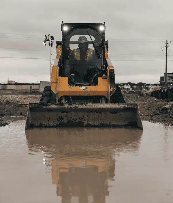 A construction worker from Professional Excavators and Construction operates a yellow skid steer loader on a muddy site, headlights shining and its reflection visible in a large puddle in front, showcasing their reliable services. - Professional Excavators & Construction