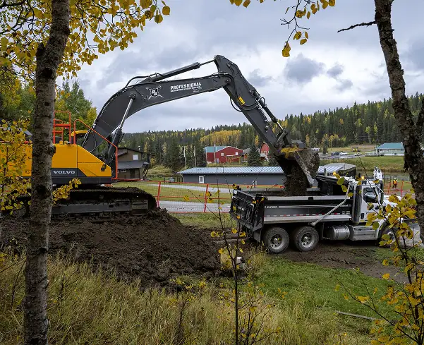 A professional excavator loads dirt into a dump truck at a construction site surrounded by autumn foliage, with buildings and a forested hillside in the background—exemplifying top-tier excavation services. - Professional Excavators & Construction