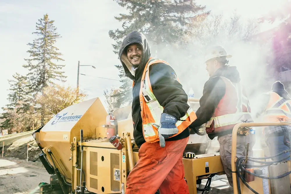 A smiling construction worker wearing a hooded jacket and orange safety vest stands by a yellow machine from Professional Excavators and Construction, with another worker in the background amid steam or dust on a sunny day. Trees are visible behind them. - Professional Excavators & Construction