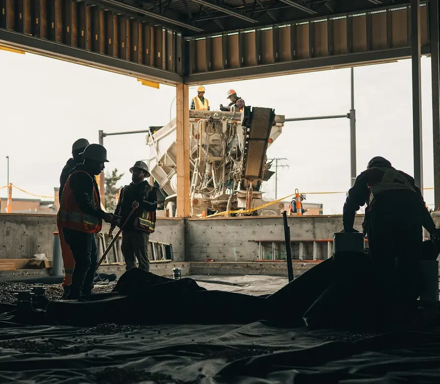 Construction workers wearing safety vests and helmets work inside a building under construction, while Professional Excavators and Construction services operate with more workers and machinery visible outside through a large open wall. - Professional Excavators & Construction