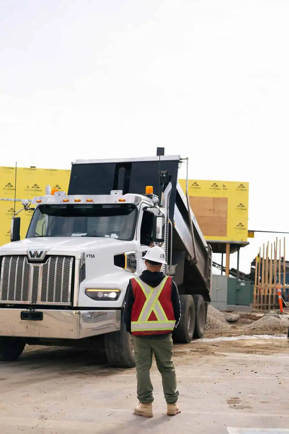 A construction worker wearing a hard hat and high-visibility vest stands in front of a large dump truck at a building site, representing the dedication of Professional Excavators and Construction services, with partially built structures in the background. - Professional Excavators & Construction